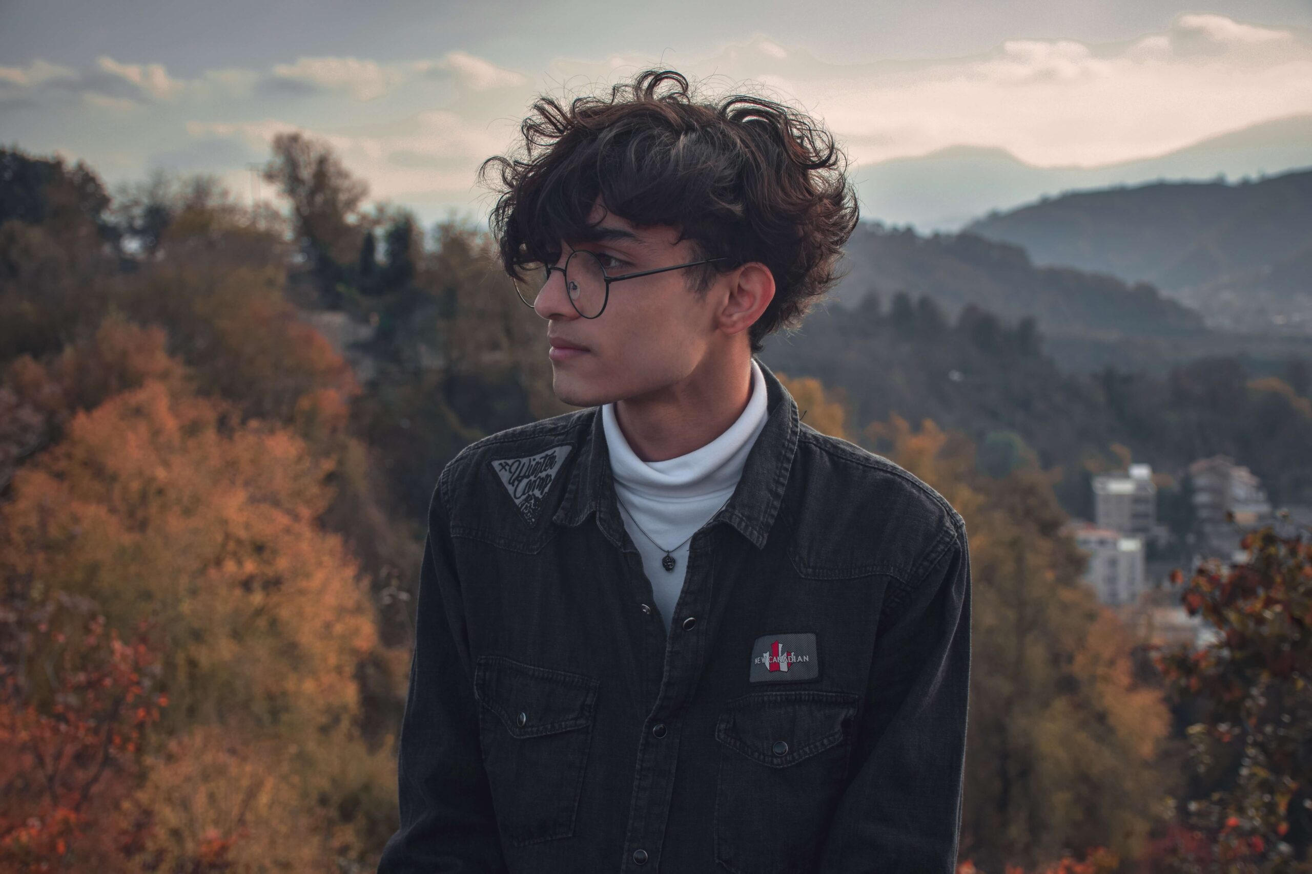 Young Man With Curly Hair and Glasses Standing Outdoors With Autumn Trees and Hills in Background.