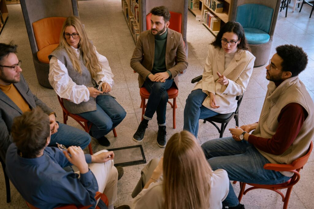 A group of adults sit on chairs in a circle in a library, engaging in a deep conversation.