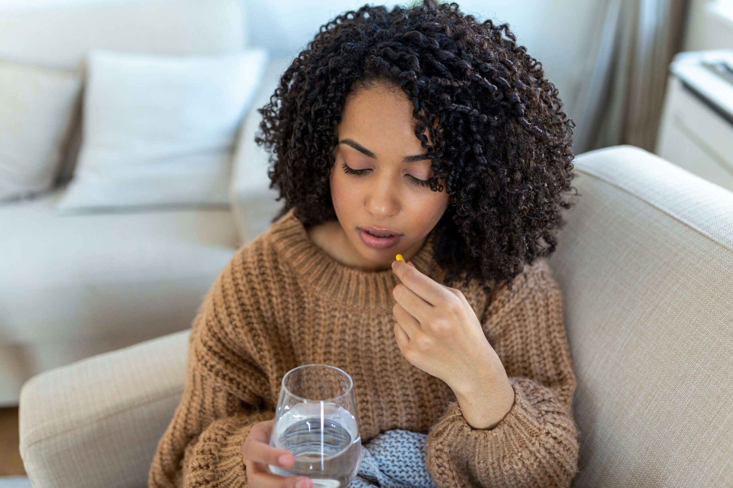 A young person sits on the couch holding a glass of water and a pill up to their mouth.