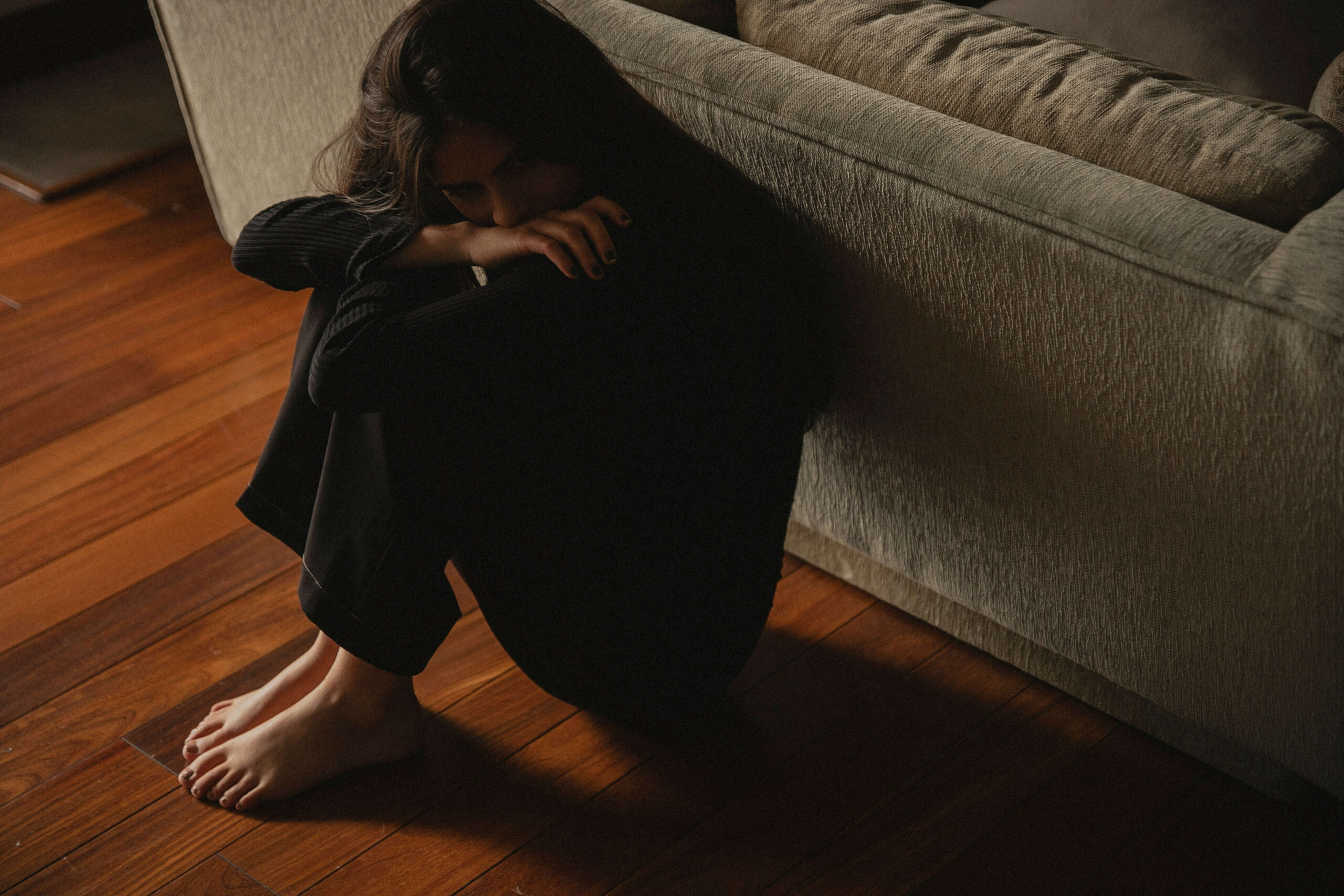 A young person sitting on the floor, leaning up against a couch with their knees pulled to their chest, appearing hopeless.