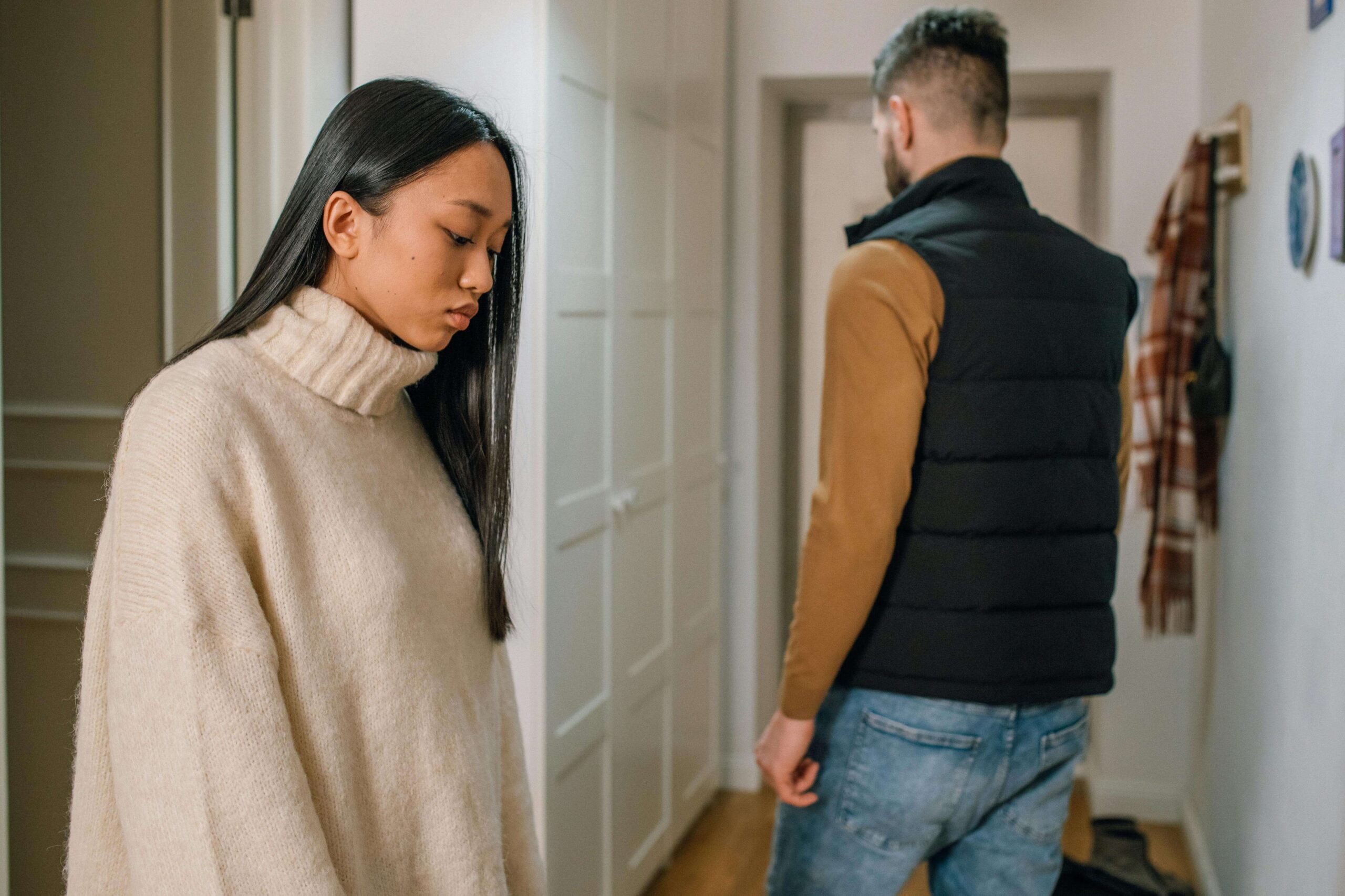 A young person with long hair looks down with a sad expression on their face, as another young person walks toward the door.