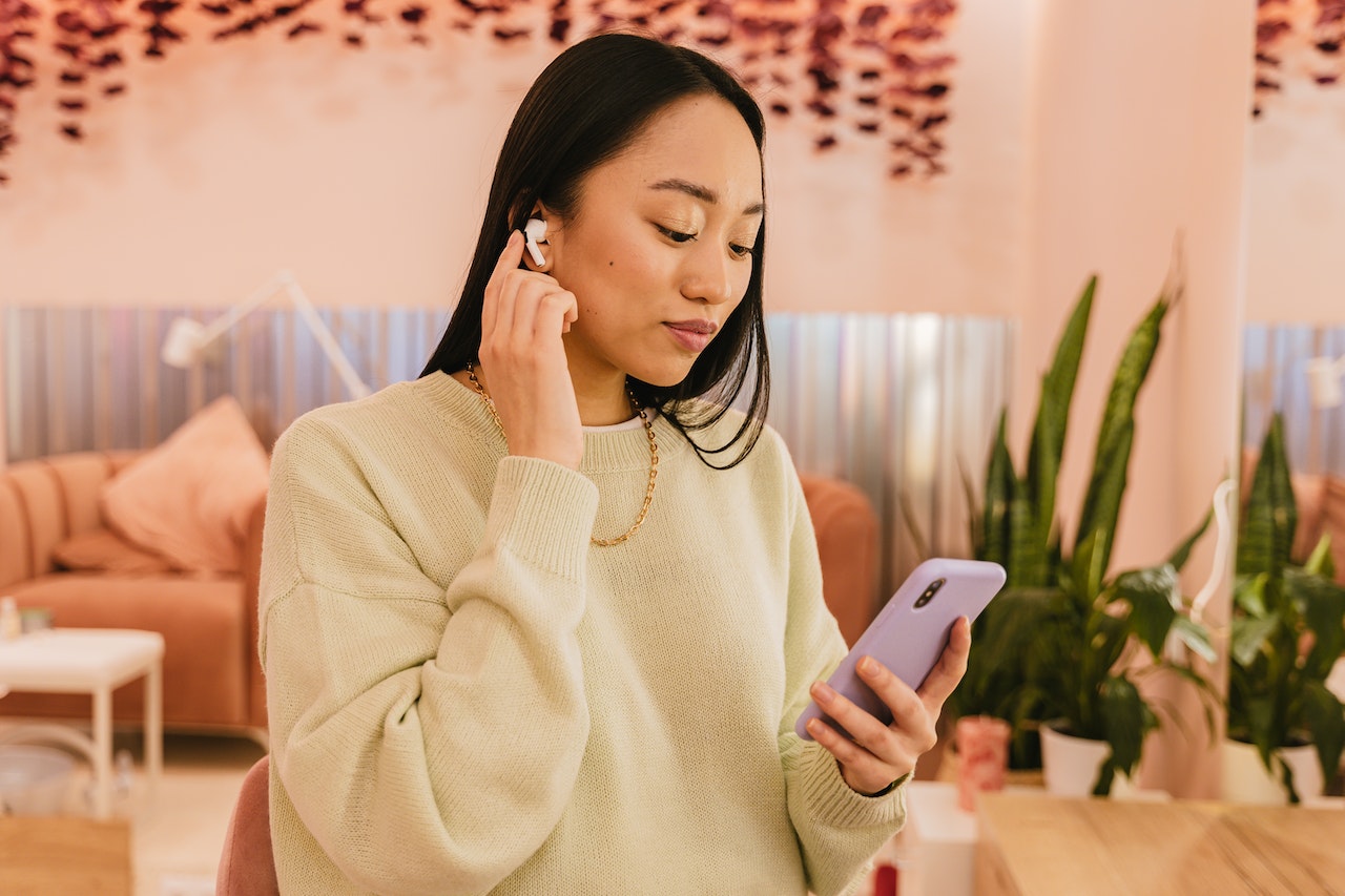 Woman Wearing Earbuds Holding a Smartphone in a Brightly Lit Room With Plants and Soft Furniture.