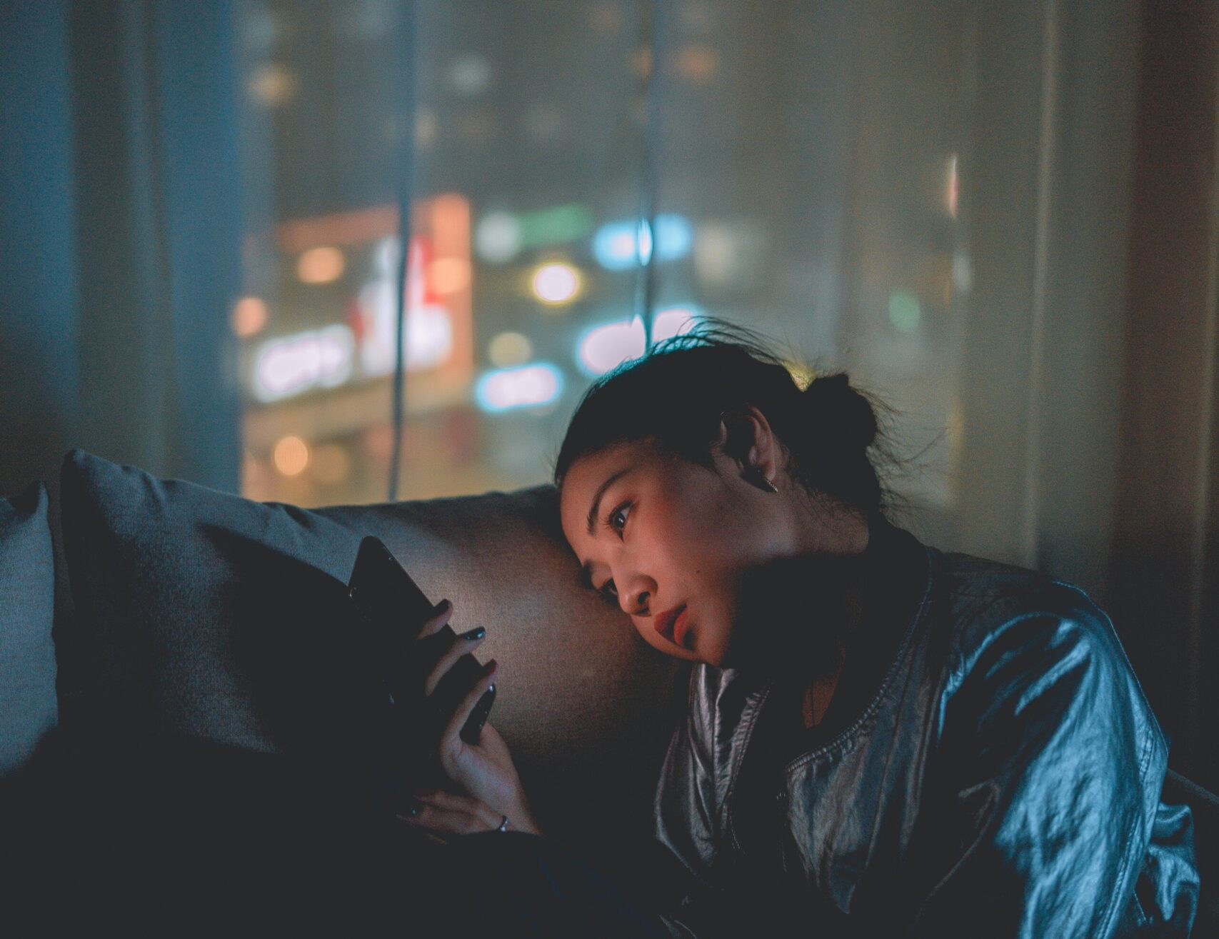 Student sitting in the dark on the couch with sad look on her face while looking at her cell phone