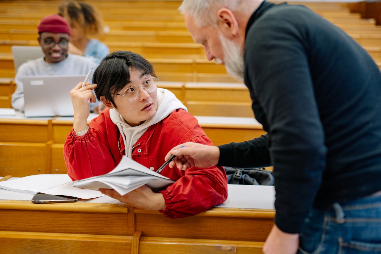 student seated in a classroom, speaking with a his professor. Professor is pointing out something in the notebook the student is holding